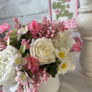 May include: A white ceramic vase with pink lettering, filled with a bouquet of pink and white flowers. The flowers include roses, daisies, and small pink blossoms. The vase is sitting on a wooden surface.