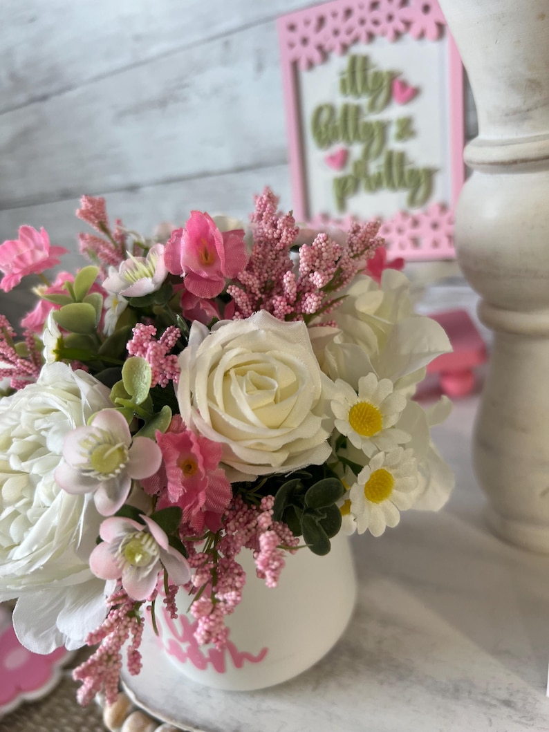 May include: A white ceramic vase with pink lettering, filled with a bouquet of pink and white flowers. The flowers include roses, daisies, and small pink blossoms. The vase is sitting on a wooden surface.