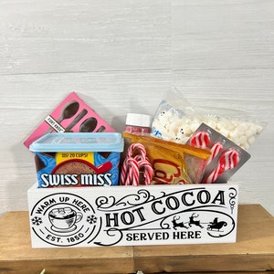 May include: White wooden box with the text "Warm Up Here Hot Cocoa Served Here" and a Santa Claus in a sleigh with reindeer. The box is filled with a variety of hot cocoa ingredients, including marshmallows, candy canes, and a box of Swiss Miss hot cocoa mix.