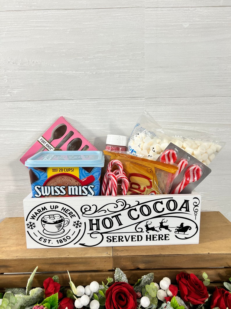 May include: White wooden box with the text "Warm Up Here Hot Cocoa Served Here" and a Santa Claus in a sleigh with reindeer. The box is filled with a variety of hot cocoa ingredients, including marshmallows, candy canes, and a box of Swiss Miss hot cocoa mix.