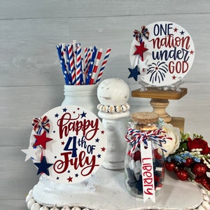 May include: A tiered tray decorated for the 4th of July with red, white, and blue decorations. The tray features a sign that says "Happy 4th of July", a jar filled with red, white, and blue stars, and a sign that says "One nation under God".
