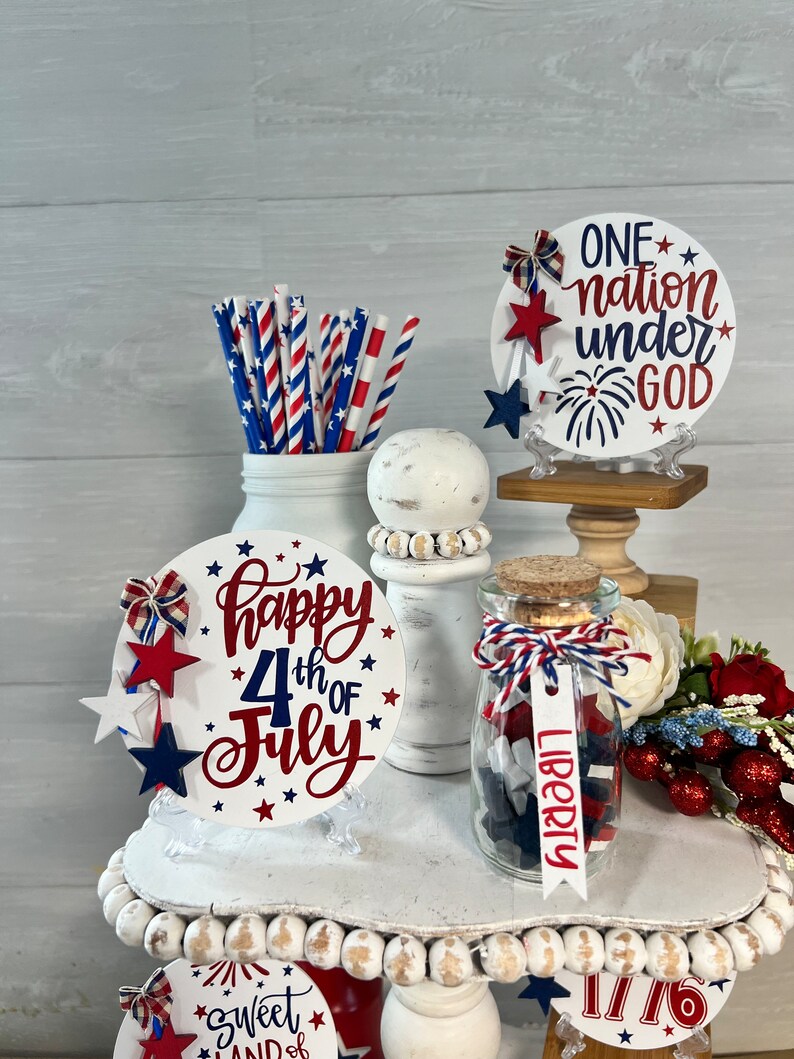 May include: A tiered tray decorated for the 4th of July with red, white, and blue decorations. The tray features a sign that says "Happy 4th of July", a jar filled with red, white, and blue stars, and a sign that says "One nation under God".