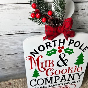 May include: White wooden sign with the text "North Pole Milk & Cookie Company Baking Santa's Favourite Since 1896". The sign is decorated with a red bow, a pine cone, and a sprig of green pine with red berries.