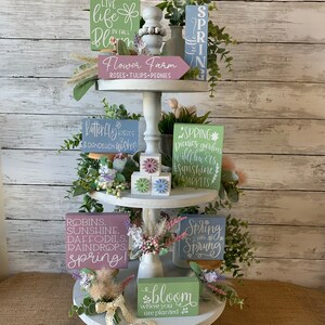 Pu&ograve; includere: Un vassoio a tre livelli decorato con cartelli a tema primaverile e accenti floreali. I cartelli presentano frasi come "Vivi la vita in fiore", "La primavera &egrave; arrivata" e "Fiorisci dove sei piantato".