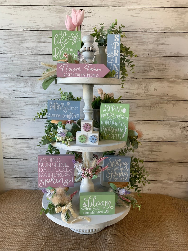 Pu&ograve; includere: Un vassoio a tre livelli decorato con cartelli a tema primaverile e accenti floreali. I cartelli presentano frasi come "Vivi la vita in fiore", "La primavera &egrave; arrivata" e "Fiorisci dove sei piantato".