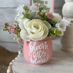 May include: A pink ceramic vase with the text "Bloom Baby Bloom" in white lettering. The vase is filled with a bouquet of white and pink flowers.