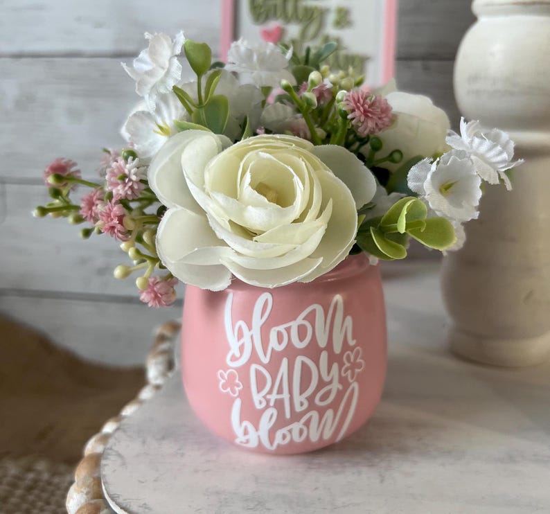 May include: A pink ceramic vase with the text "Bloom Baby Bloom" in white lettering. The vase is filled with a bouquet of white and pink flowers.