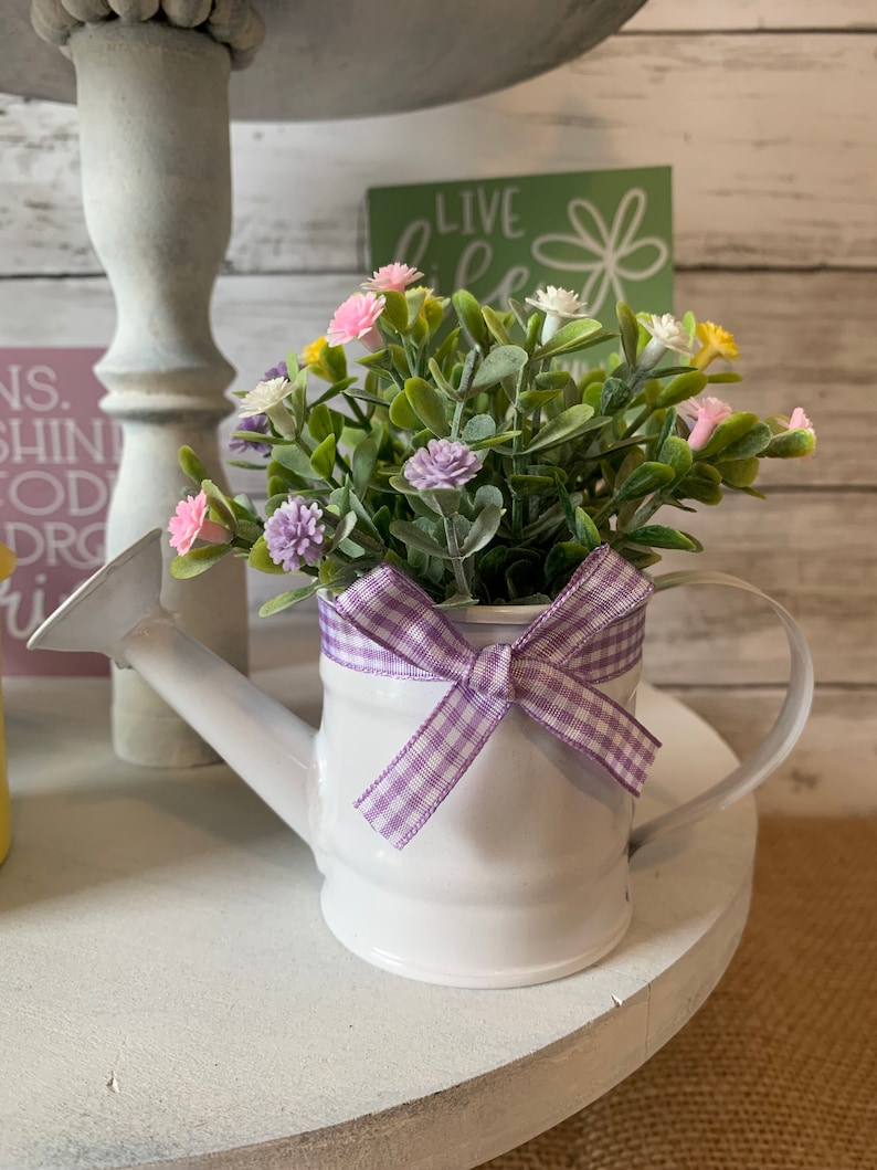 May include: A white metal watering can filled with greenery and colorful flowers. A purple and white gingham ribbon is tied around the can. The watering can is sitting on a white, tiered stand.