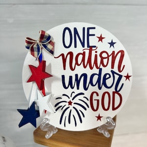 May include: A white round wooden sign with the text "ONE nation under GOD" in red, blue, and white lettering. The sign is decorated with red, white, and blue stars and a red, white, and blue bow. The sign is sitting on a wooden stand.