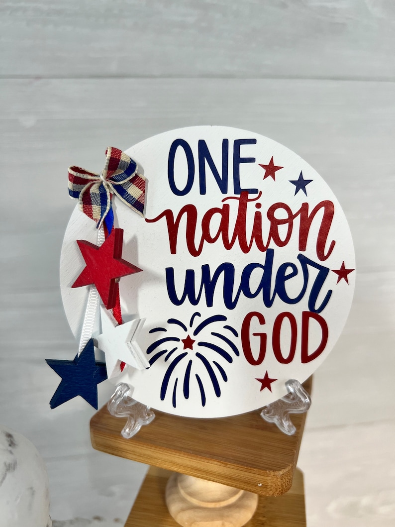 May include: A white round wooden sign with the text "ONE nation under GOD" in red, blue, and white lettering. The sign is decorated with red, white, and blue stars and a red, white, and blue bow. The sign is sitting on a wooden stand.