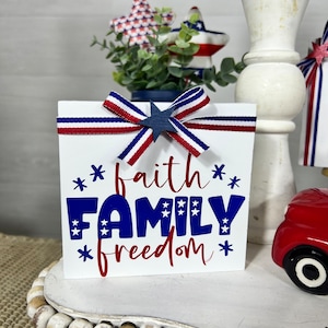 May include: A white wooden block with the words "faith family freedom" in red and blue lettering. The block is decorated with a red, white, and blue ribbon bow and a blue star.