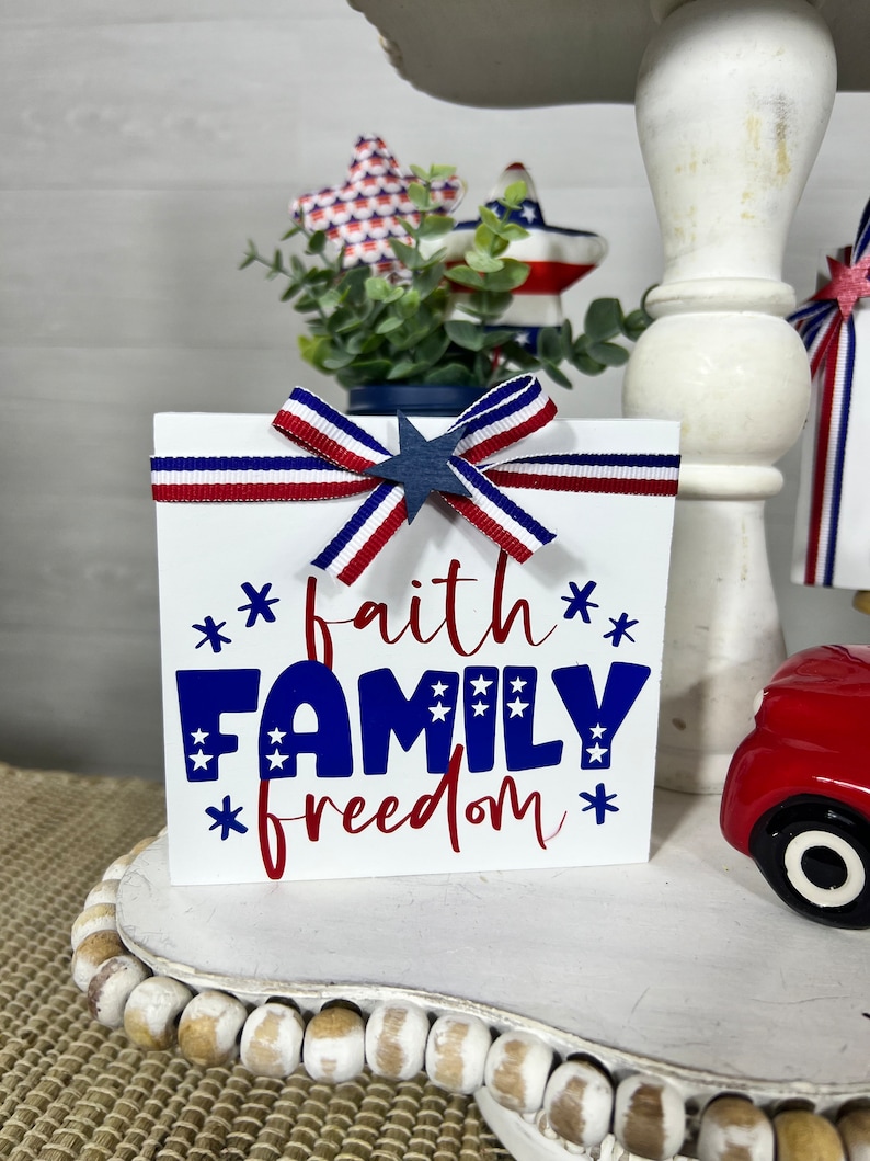 May include: A white wooden block with the words "faith family freedom" in red and blue lettering. The block is decorated with a red, white, and blue ribbon bow and a blue star.