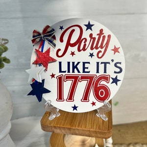 May include: A white round sign with red and blue text that says "Party Like It's 1776" with red and blue stars around the text. The sign is on a small wooden stand.