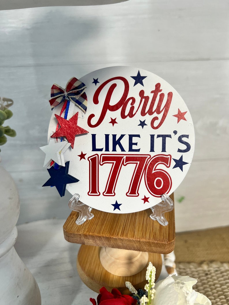 May include: A white round sign with red and blue text that says "Party Like It's 1776" with red and blue stars around the text. The sign is on a small wooden stand.