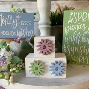 Pu&ograve; includere: Tre blocchi di legno impilati uno sopra l'altro, ogni blocco con un ritaglio di fiore di colore diverso sul davanti. Il blocco superiore ha un fiore rosa, il blocco centrale ha un fiore verde e il blocco inferiore ha un fiore blu. I blocchi sono posizionati su un vassoio a pi&ugrave; livelli bianco con altre decorazioni a tema primaverile.