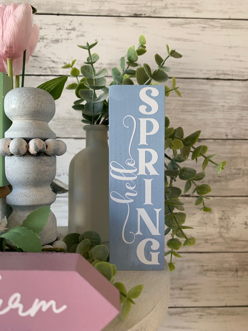 Pu&ograve; includere: Un cartello in legno blu chiaro con la parola "SPRING" scritta verticalmente in lettere bianche. Il cartello &egrave; decorato con un "hello" bianco in corsivo nell'angolo in alto a sinistra. Il cartello &egrave; appoggiato su una superficie bianca con piante verdi sullo sfondo.
