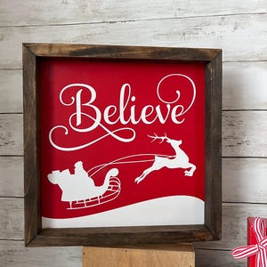 May include: A red wooden sign with a white frame. The sign says "Believe" in a cursive font. There is a white silhouette of Santa Claus in a sleigh pulled by a reindeer.