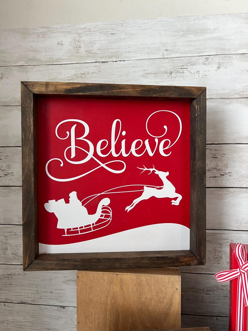 May include: A red wooden sign with a white frame. The sign says "Believe" in a cursive font. There is a white silhouette of Santa Claus in a sleigh pulled by a reindeer.