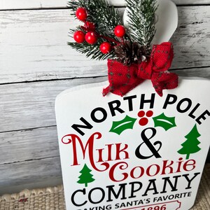 May include: White wooden sign with the text "North Pole Milk & Cookie Company Baking Santa's Favourite Since 1896". The sign is decorated with a red bow, a pine cone, and a sprig of green pine with red berries.
