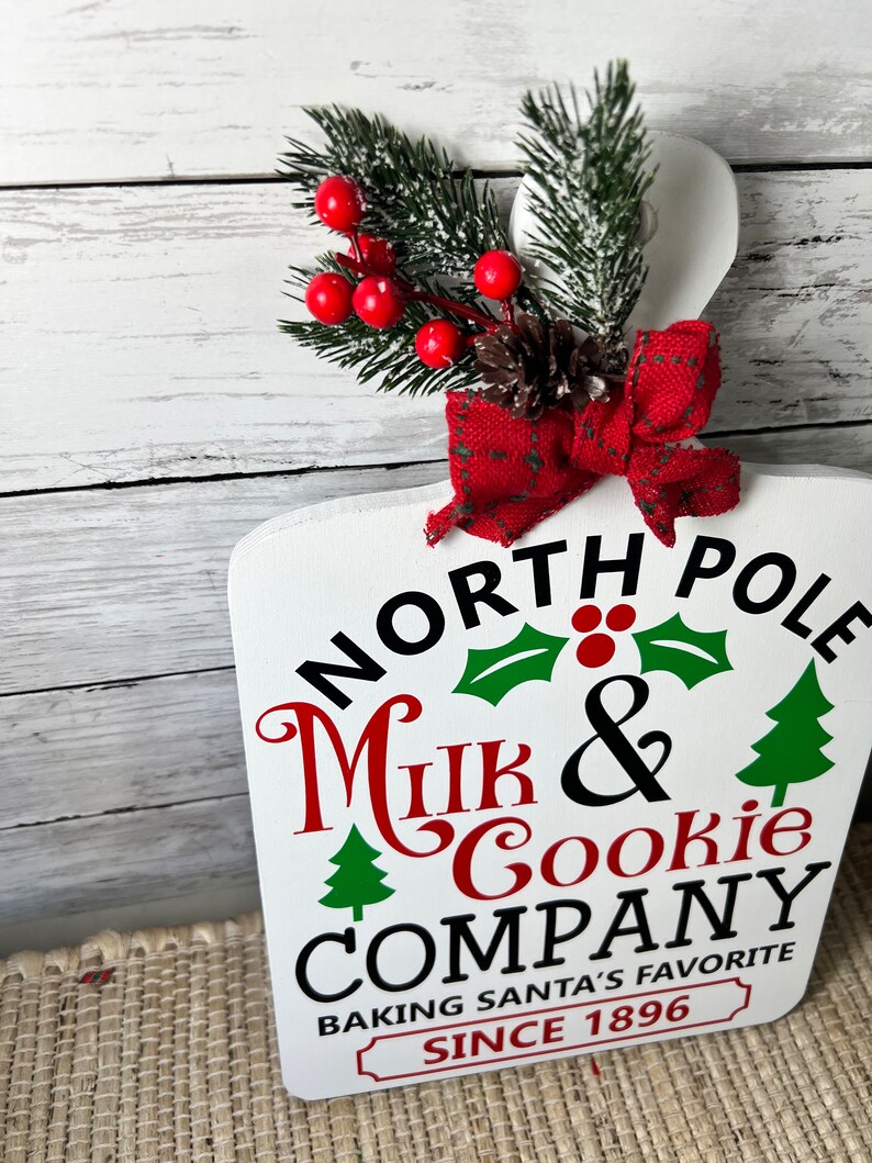 May include: White wooden sign with the text "North Pole Milk & Cookie Company Baking Santa's Favourite Since 1896". The sign is decorated with a red bow, a pine cone, and a sprig of green pine with red berries.