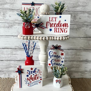 Pode incluir: Uma bandeja de n&iacute;veis decorada com decora&ccedil;&atilde;o patri&oacute;tica vermelha, branca e azul. A bandeja apresenta placas com o texto "Let freedom ring", "My home sweet home" e "Red white and blessed".