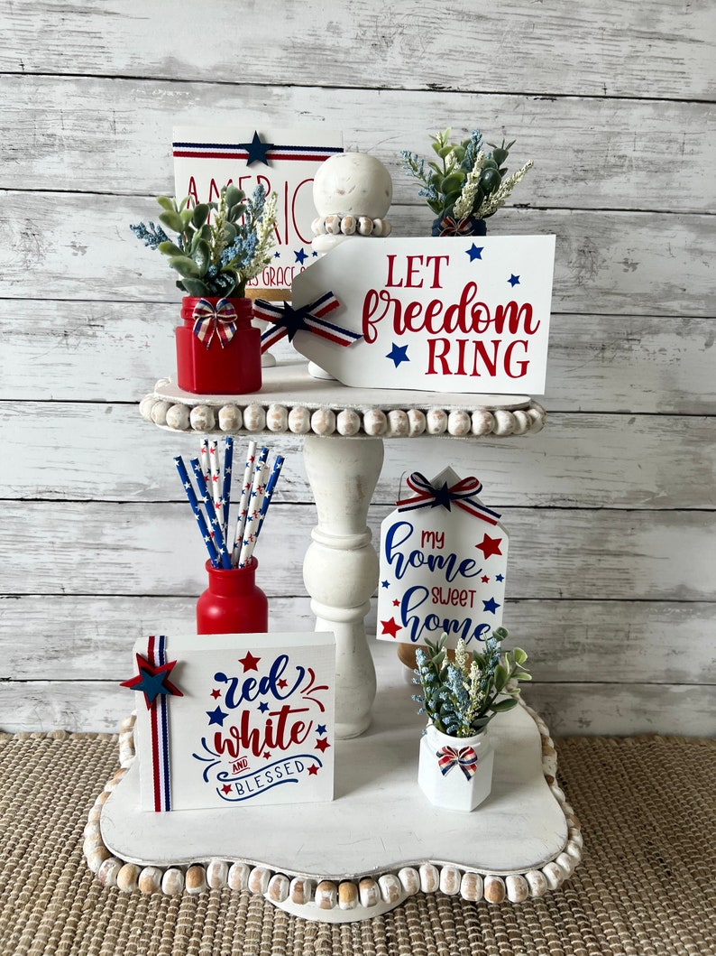 Pode incluir: Uma bandeja de n&iacute;veis decorada com decora&ccedil;&atilde;o patri&oacute;tica vermelha, branca e azul. A bandeja apresenta placas com o texto "Let freedom ring", "My home sweet home" e "Red white and blessed".