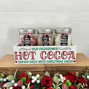 May include: A white wooden box with the text "Old Fashioned Hot Cocoa Served Daily With Christmas Cheer" on the front. The box contains three glass jars filled with candy canes, hot cocoa mix, and marshmallows. The jars are decorated with red and white twine and black chalkboard tags.
