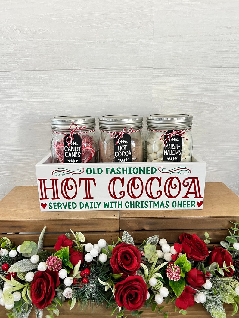 May include: A white wooden box with the text "Old Fashioned Hot Cocoa Served Daily With Christmas Cheer" on the front. The box contains three glass jars filled with candy canes, hot cocoa mix, and marshmallows. The jars are decorated with red and white twine and black chalkboard tags.