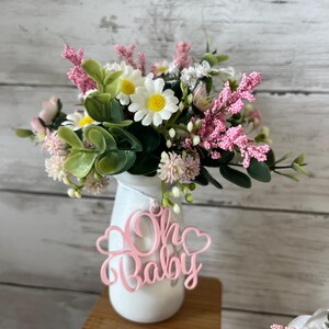 May include: A white vase with a pink "Oh Baby" sign hanging from it. The vase is filled with a bouquet of pink and white flowers.