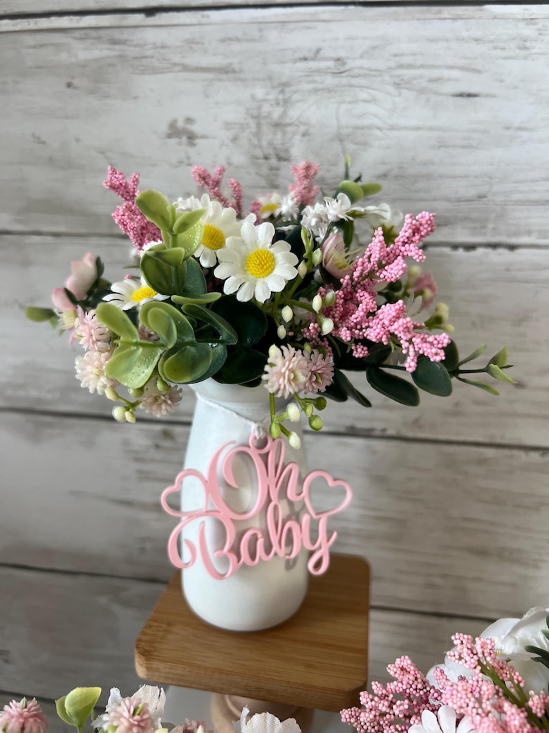 May include: A white vase with a pink "Oh Baby" sign hanging from it. The vase is filled with a bouquet of pink and white flowers.
