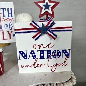May include: White wooden sign with red, white, and blue ribbon and a star. The sign says "one nation under God" in blue and red lettering with stars.