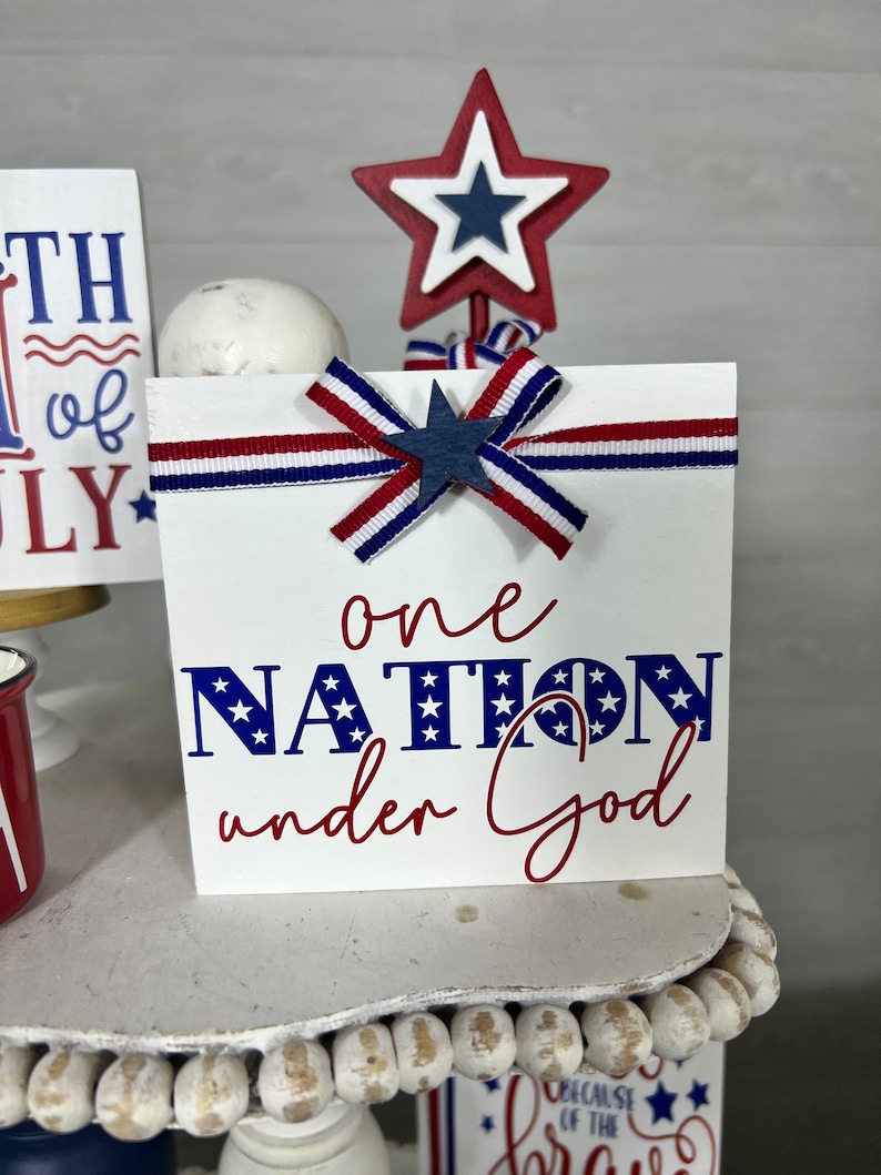 May include: White wooden sign with red, white, and blue ribbon and a star. The sign says "one nation under God" in blue and red lettering with stars.