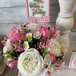 May include: A white ceramic vase filled with a bouquet of pink and white flowers. The flowers include roses, daisies, and sprigs of pink flowers. The vase is sitting on a wooden surface with a pink sign that says "itty bitty & pretty".