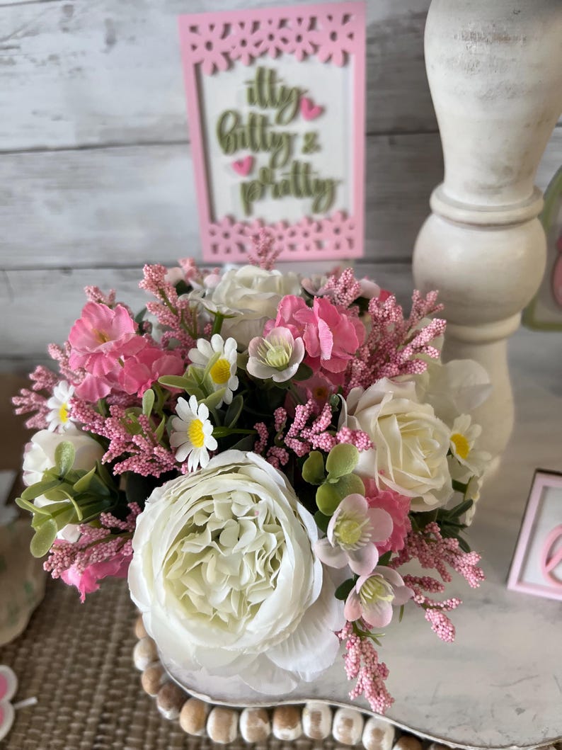 May include: A white ceramic vase filled with a bouquet of pink and white flowers. The flowers include roses, daisies, and sprigs of pink flowers. The vase is sitting on a wooden surface with a pink sign that says "itty bitty & pretty".