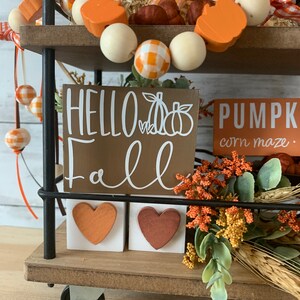 May include: A black metal tiered stand with three shelves decorated for fall. The top shelf has a plaid ribbon, orange and white wooden beads, and orange pumpkin-shaped beads. The middle shelf has a brown wooden sign that says "HELLO fall" with a white pumpkin graphic. The bottom shelf has two white wooden blocks with orange heart-shaped decorations. The stand is on a wooden surface with a bouquet of orange and green fall foliage.