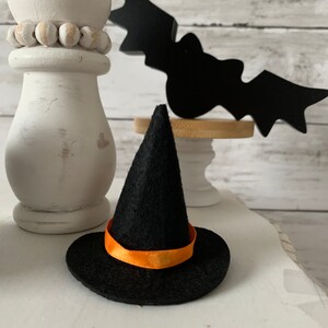 May include: A small black witch hat with an orange ribbon. The hat is sitting on a white surface with a white pillar and a black bat cutout in the background.