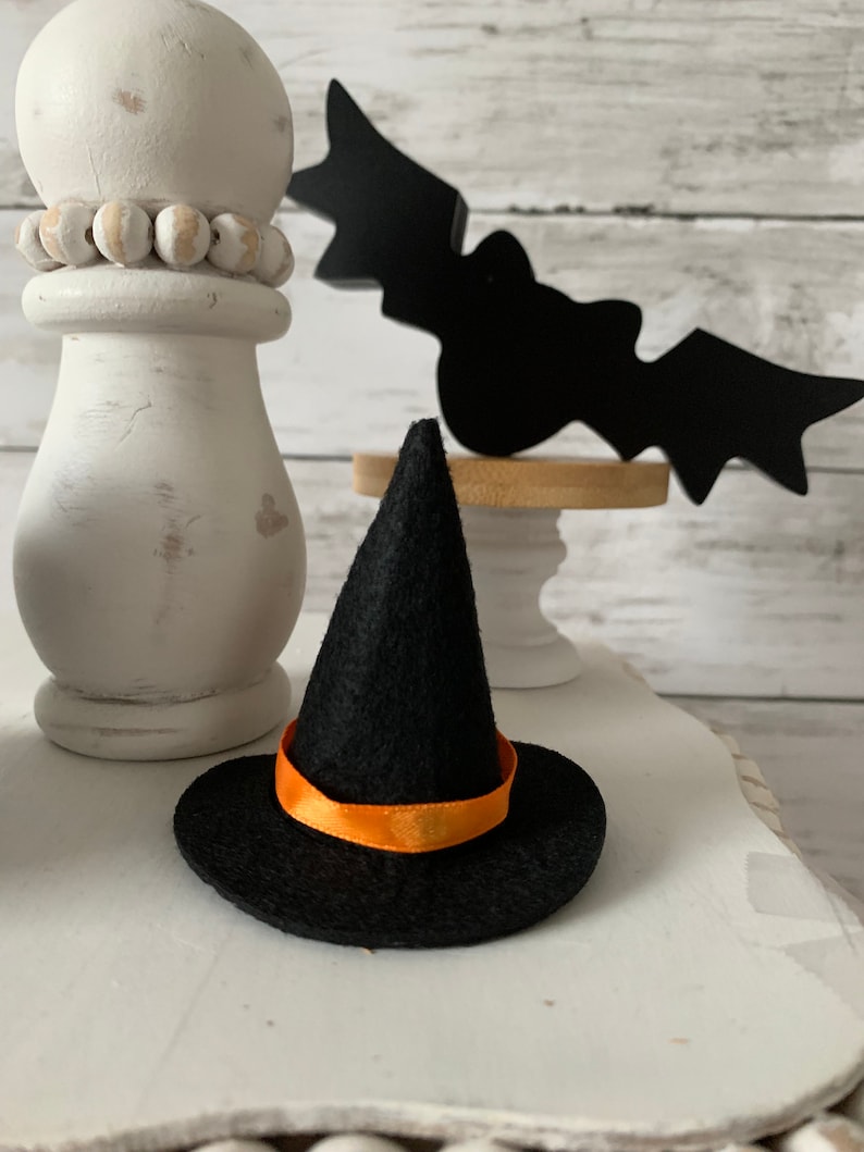 May include: A small black witch hat with an orange ribbon. The hat is sitting on a white surface with a white pillar and a black bat cutout in the background.
