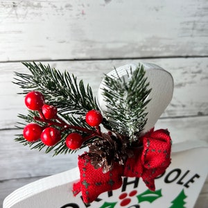 May include: White wooden sign with the text "North Pole Milk & Cookie Delivery" in black lettering. The sign is decorated with a red bow, a pine sprig with red berries, and a brown pine cone.