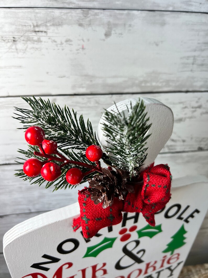 May include: White wooden sign with the text "North Pole Milk & Cookie Delivery" in black lettering. The sign is decorated with a red bow, a pine sprig with red berries, and a brown pine cone.