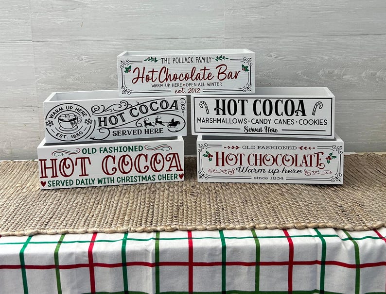 May include: Four white wooden boxes stacked on top of each other. Each box has a different design with the words "Hot Cocoa" or "Hot Chocolate" in red and green lettering. The top box says "The Pollack Family Hot Chocolate Bar Warm Up Here Open All Winter est 2012". The second box says "Warm Up Here Hot Cocoa Served Here Est 1850". The third box says "Hot Cocoa Marshmallows Candy Canes Cookies Served Here". The bottom box says "Old Fashioned Hot Chocolate Warm Up Here Since 1854".