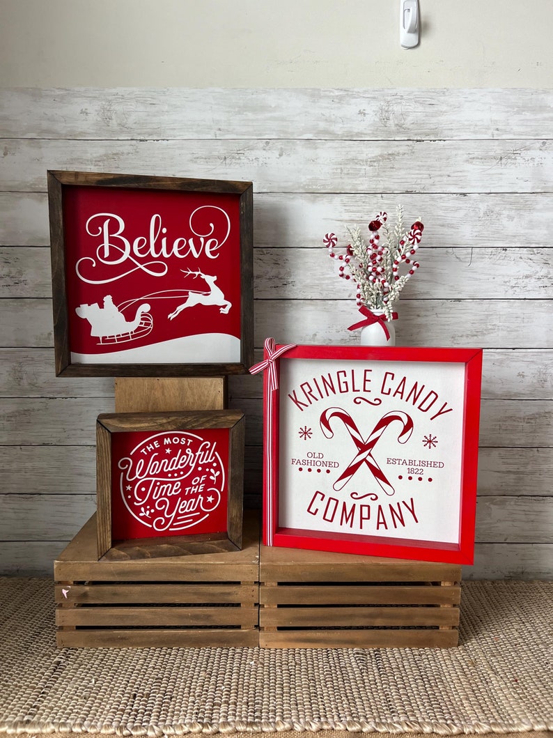 May include: Three wooden signs with red backgrounds and white lettering. The signs feature Christmas-themed sayings: "Believe", "The Most Wonderful Time of the Year", and "Kringle Candy Company". The signs are displayed on wooden crates against a white wood background.