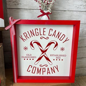 May include: Red wooden framed sign with white background and red text that reads "Kringle Candy Company" with a red candy cane graphic and the text "Old Fashioned" and "Established 1822".