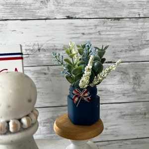 Pode incluir: Um pequeno frasco azul com uma fita vermelha, branca e azul amarrada em volta da parte superior. O frasco est&aacute; cheio de um buqu&ecirc; de flores brancas e verdes. O frasco est&aacute; sentado em um pequeno pedestal de madeira.