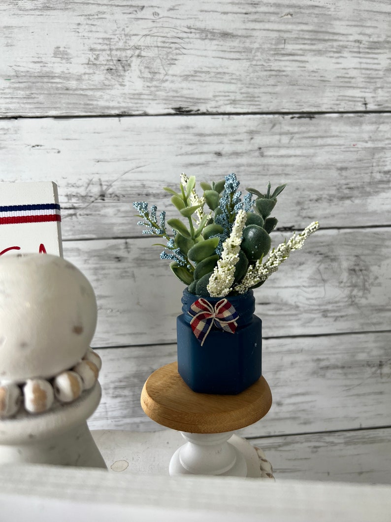 Pode incluir: Um pequeno frasco azul com uma fita vermelha, branca e azul amarrada em volta da parte superior. O frasco est&aacute; cheio de um buqu&ecirc; de flores brancas e verdes. O frasco est&aacute; sentado em um pequeno pedestal de madeira.