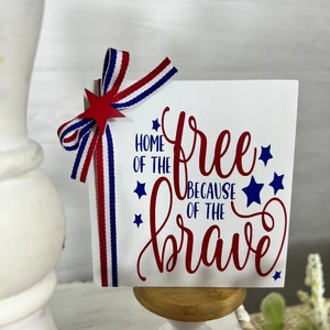 May include: A white wooden sign with red and blue stars and the text "Home of the free because of the brave". The sign is decorated with a red, white, and blue ribbon bow.