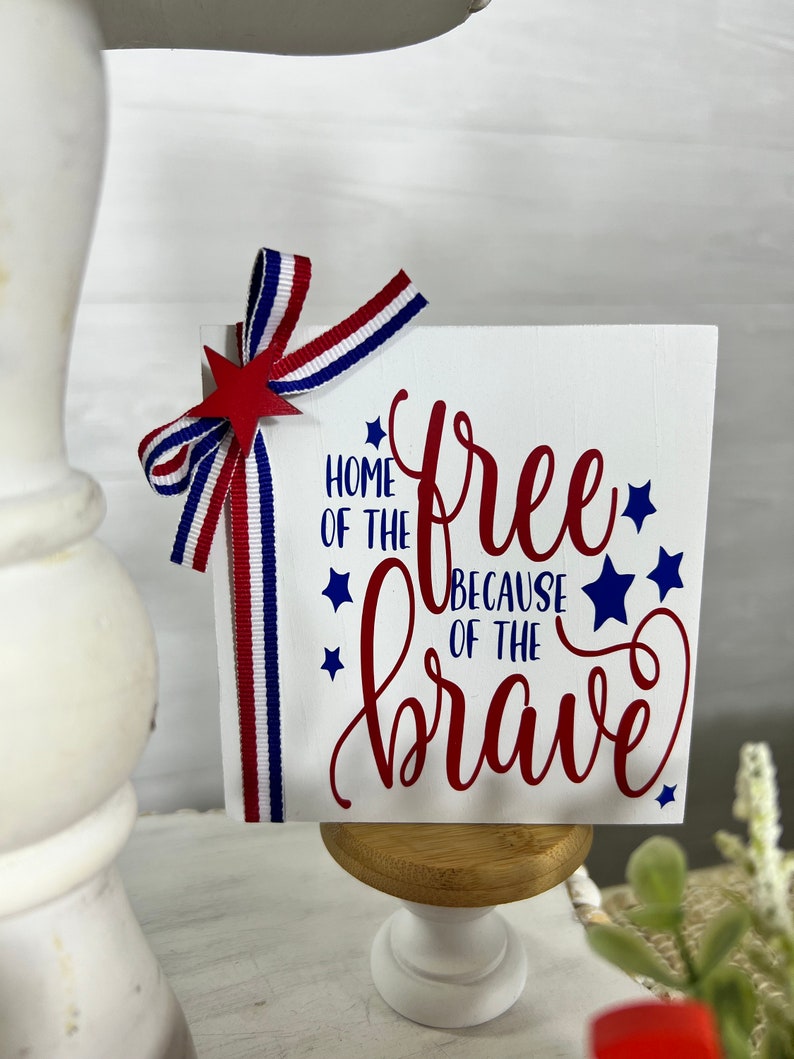 May include: A white wooden sign with red and blue stars and the text "Home of the free because of the brave". The sign is decorated with a red, white, and blue ribbon bow.