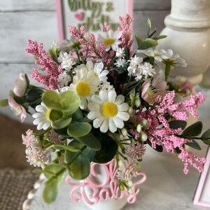 May include: A white vase with a pink "Oh Baby" sign in front of it. The vase is filled with a bouquet of pink and white flowers, including daisies and baby's breath.