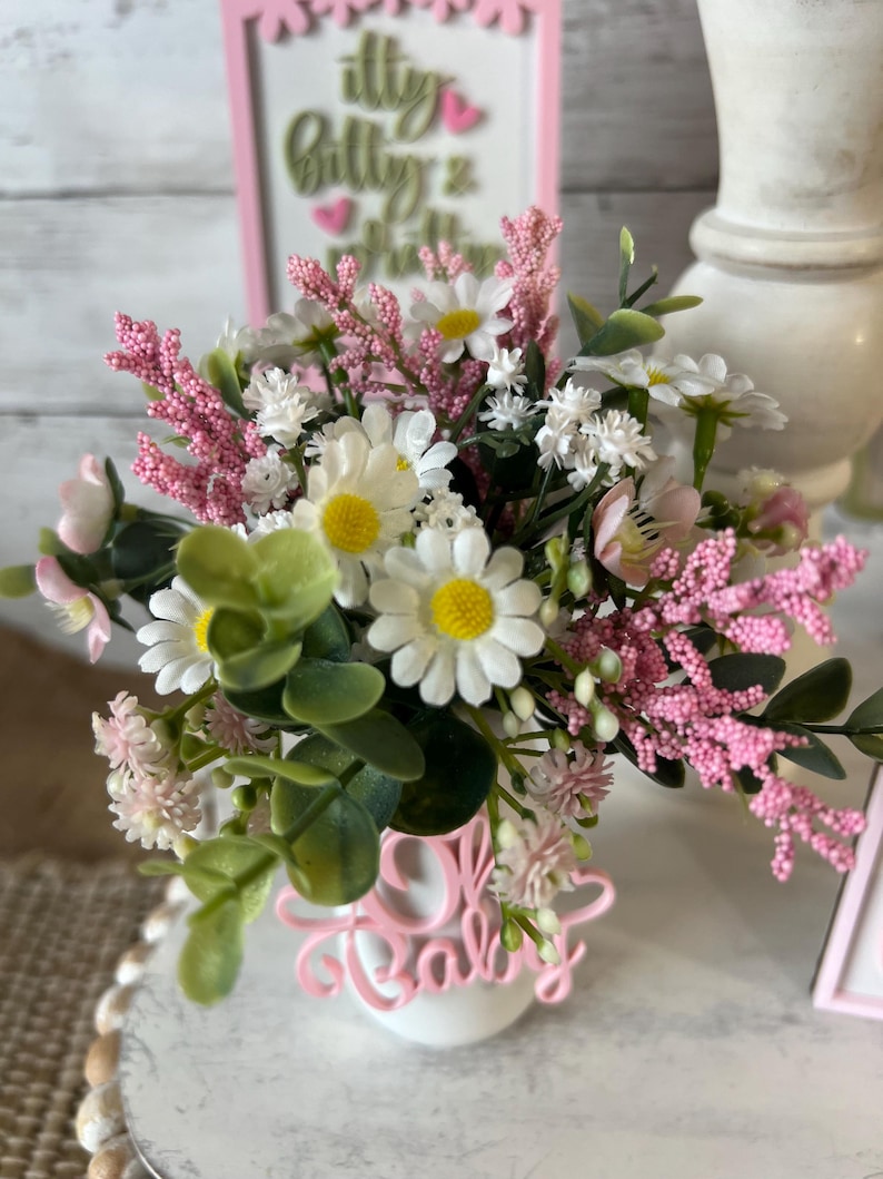 May include: A white vase with a pink "Oh Baby" sign in front of it. The vase is filled with a bouquet of pink and white flowers, including daisies and baby's breath.