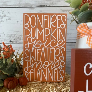 May include: A wooden sign with the text "Bonfires, Pumpkin Spice, Sweaters, Smores & Flannels" in white lettering on an orange background.