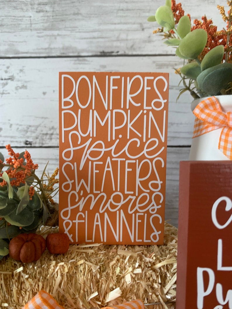 May include: A wooden sign with the text "Bonfires, Pumpkin Spice, Sweaters, Smores & Flannels" in white lettering on an orange background.